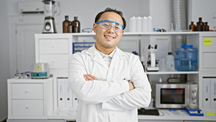 Naklejka na ściany i meble Smiling, confident young chinese man, a pro in science, at his lab station, standing with iconic arms crossed in the epicenter of research and medicine.