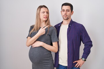 Young couple expecting a baby standing over white background pointing aside worried and nervous...