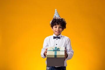 Child boy with birthday cap on head holding giftbox over yellow background. Birthday and...