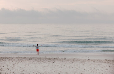 Kleiner Junge mit roten Hosen und blauem Shirt ist alleine am Strand die Hände ausgestreckt schaut auf das Meer und spielt mit den Wellen