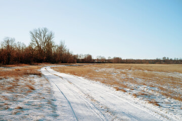 Snow-covered road among dry yellow grass in a spring field on a sunny day. Autumn or spring background.