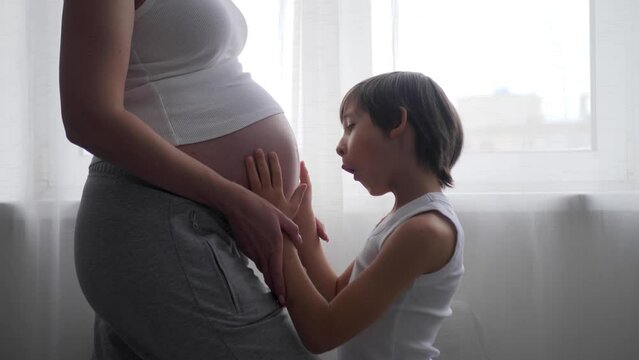 A pregnant woman in a white T-shirt stands at the window with a curtain, her eldest son of six years old. Boy strokes, kisses, hugs the belly of a pregnant mother.