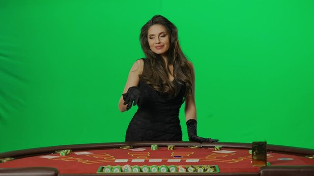 Portrait of female on chroma key green screen. Woman in dress stands at the blackjack poker table pointing at camera showing table. Slow motion.