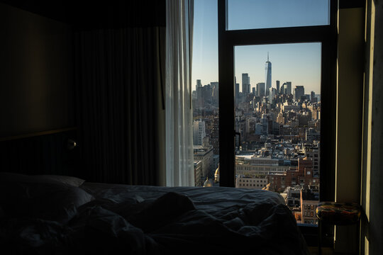 A view of the Manhattan skyline from a midtown New York City hotel bedroom, including views of One World Trade Center.  Sunny day with blue skies, and an unmade bed in the foreground.