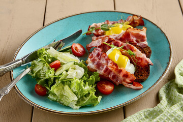 Close-up side view of a poached egg on toast with crispy bacon, accompanied by a fresh salad on a blue plate