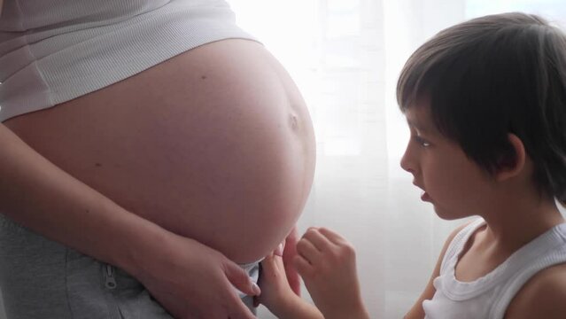 A pregnant woman in a white T-shirt stands at the window with a curtain, her eldest son of six years old. Boy strokes, kisses, hugs the belly of a pregnant mother.