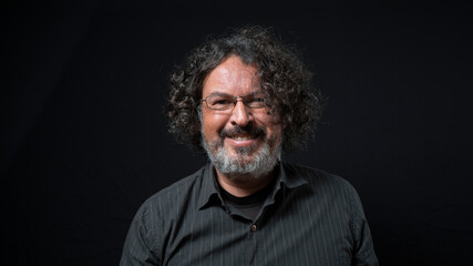 Man with white beard and black curly hair with happy expression, seen from front, wearing black shirt against black background