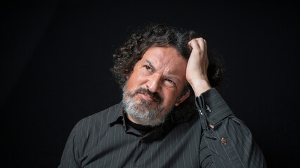 Man with white beard and black curly hair with confused expression, scratching head with his hand, wearing black shirt against black background