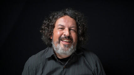 Man with white beard and black curly hair with happy expression, with half closed eyes, wearing black shirt against black background