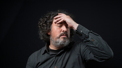 Man with white beard and black curly hair with worried expression, with eyes closed and hand on forehead, wearing black shirt against black background
