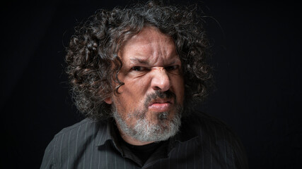 Man with white beard and black curly hair with displeased expression, looking towards camera, wearing black shirt against black background