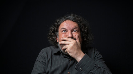 Man with white beard and black curly hair with funny expression, covering his mouth with hand, wearing black shirt against black background