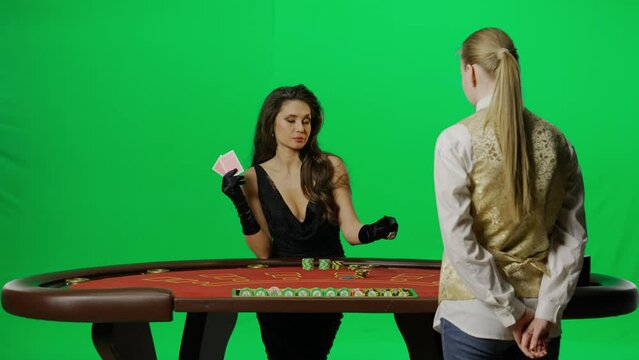 Portrait of elegant female on chroma key green screen. Attractive woman at blackjack poker table with croupier, holding cards placing bet smiling.