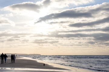 This image encapsulates the peaceful experience of a beach walk under a vast, expressive sky. A small group of people and a solitary dog on the beach create a sense of scale and companionship, against