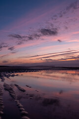 The image is a symphony of colors as twilight descends upon a still beach. The calm water reflects a tapestry of purples and pinks from the sky, where clouds are painted with the soft, fading light of