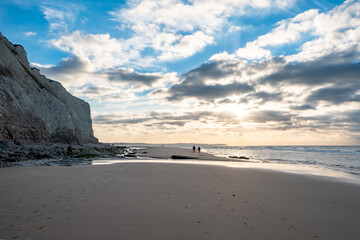 This photograph captures the grandeur of a coastal landscape at sunset. Two distant figures are seen walking near the water's edge, providing a sense of scale against the imposing cliff face. The sun