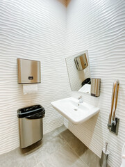 A toilet in the hotel, a sink with a mirror, a metal trash can, a paper towel is served from a vending machine.