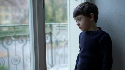 introspective kid standing by window gazes out from apartment with sad lonely expression and...
