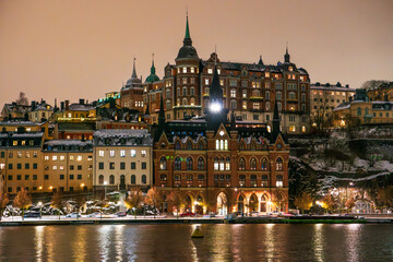 Fototapeta premium Stockholm, district of Sodermalm at night in winter with snow on the ground, historic buildings. Cloudy sky, lake reflections.