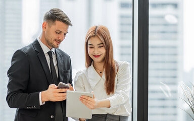 Two diverse smart businessman, woman wearing formal suits, talking, working, discussing, planning their project together, standing beside window, looking at tablet. Business Concept.