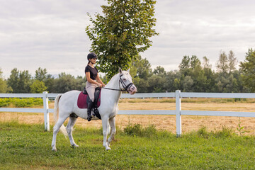 Pretty girl, a horseback rider riding beautiful snow white horse, walking along a path on a sunny...