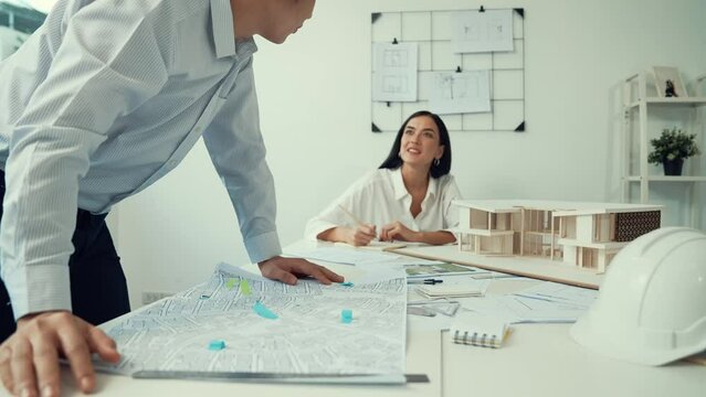 Skilled caucasian interior designer working with house model while discussing with asian architect engineer about house plan on meeting table with blueprint and house model scatter around. Immaculate.