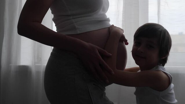 A pregnant woman in a white T-shirt stands at the window with a curtain, her eldest son of six years old. Boy strokes, kisses, hugs the belly of a pregnant mother.