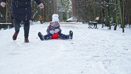Fototapeta na wymiar Happy Caucasian Girl Kid Having Fun Playing on Snow With Her Mother Pulling Her on Plastic Snow Slide Drag Sled Down Alley in City Park