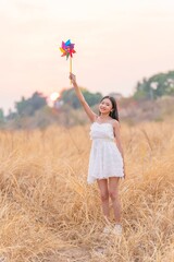 Girl in White Long Dress Holding a Colorful Windmill Toy in a Dry Grass Meadow