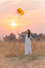 Beautiful girl with balloons in the field. Carefree Serenity: Girl in White Long Dress Embracing Freedom, Holding Balloons in a Dry Grass Meadow