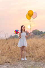 Girl in White Dress Holding Yellow Balloons and Colorful Windmill Toy in a Dry Grass Meadow