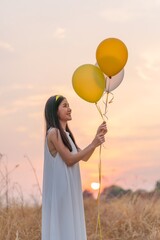 Beautiful girl with balloons in the field. Carefree Serenity: Girl in White Long Dress Embracing Freedom, Holding Yellow Balloons in a Dry Grass Meadow