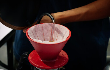 Hands of barista brewing a drip hot espresso by pouring hot water into red cup with filter paper drip coffee from kettle, close up.