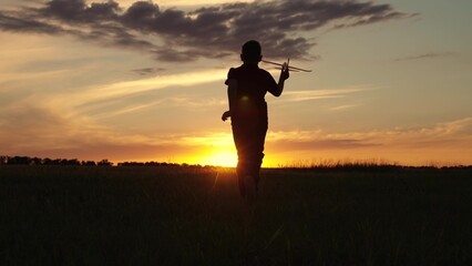 Happy kid runs with toy airplane against background of summer nature in field at sunset. Silhouette...