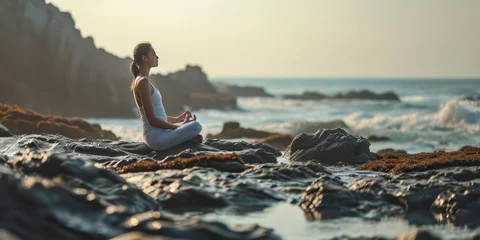 Zelfklevend Fotobehang A fitness enthusiast, a young woman, finds serenity in her yoga practice, savoring the gentle caress of the wind and inhaling the crisp sea breeze as she balances on the rocky beach © Nattadesh