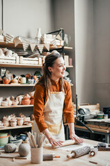 Artisan woman in lighted pottery studio, rolling out clay for modelling. Pottery studio with a variety of ceramics and a focused artisan