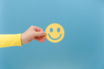Close up of female hand holding yellow happy smile face, isolated over blue background with copy...