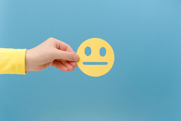 Close up of female hand holding yellow emoticon with boring face, posing isolated over plain blue...