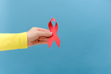 Close up of female hand holding red ribbon awareness, isolated on blue studio background. World AIDS day in December month campaign. Symbol of awareness and support for people living w HIV and STD