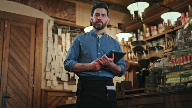 Front view of handsome caucasian man wearing black apron using digital tablet while standing near bar counter. Competent male bartender smiling and looking at camera during working day shift.