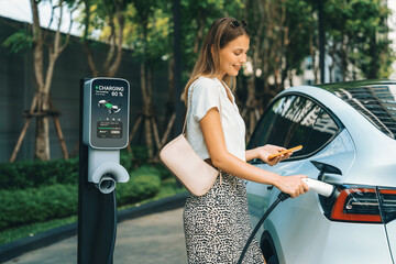 Young woman travel with EV electric car charging in green sustainable city outdoor garden in...