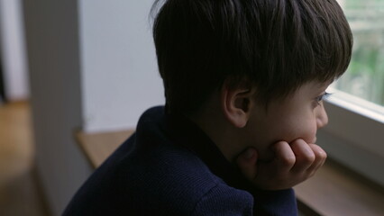 Thoughtful young boy staring at view from apartment window with hand in chin, pensive child with daydreaming expression