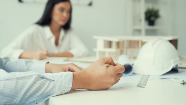 Professional architect engineer drawing blueprint and working together with young beautiful caucasian coworker on meeting table with safety helmet, house model, blueprint scatter around. Immaculate.