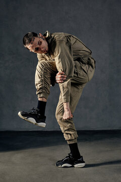 Strong male with tattooed body and face, earrings, beard. Dressed in khaki jumpsuit and black sneakers. Dancing on gray background. Dancehall, hip-hop