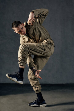Strong guy with tattooed body and face, earrings, beard. Dressed in khaki overalls and black sneakers. Dancing on gray background. Dancehall, hip-hop