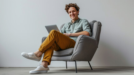 Young Caucasian man with curly hair, comfortably seated in a modern gray armchair, smiling and relaxed while using a laptop on his lap - obrazy, fototapety, plakaty