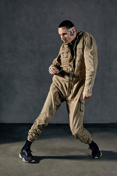 Active performer with tattooed body, earrings, beard. Dressed in khaki overalls and black sneakers. Dancing on gray background. Dancehall, hip-hop