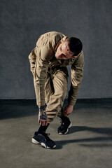 Flexible male with tattooed body and face, earrings, beard. Dressed in khaki jumpsuit, black sneakers. Dancing on gray background. Dancehall, hip-hop