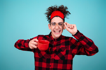 Naklejka premium Portrait of a cute guy who woke up in the morning holding a red coffee mug in his hands. Blue background. A man in a red-checked shirt.