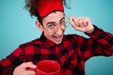 Portrait of a cute guy who woke up in the morning holding a red coffee mug in his hands. Blue background. A man in a red-checked shirt. - obrazy, fototapety, plakaty